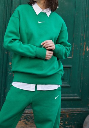 Personne portant un sweat-shirt et un pantalon de jogging verts assortis Nike avec une chemise à col blanc en dessous, debout devant une porte verte usée.