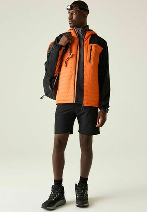 Mann trägt eine orange-schwarze Jacke, schwarze Shorts, Wanderschuhe, einen Rucksack und eine Stirnlampe und schaut nach links.