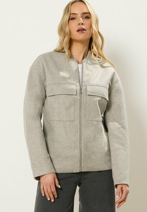 Nainen, joka käyttää vaaleanharmaata vetoketjullista takkia, jossa on kaksi rintataskun läppää ja pitkät hihat, yhdistetty tummanharmaisiin housuihin.