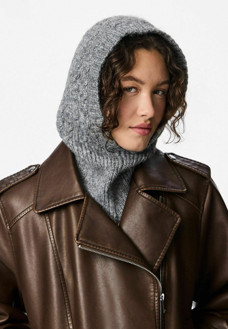 Cappuccio grigio lavorato a maglia con collo alto, abbinato a una giacca di pelle marrone scuro con colletto a punte e chiusura a zip. I materiali strutturati creano un contrasto.