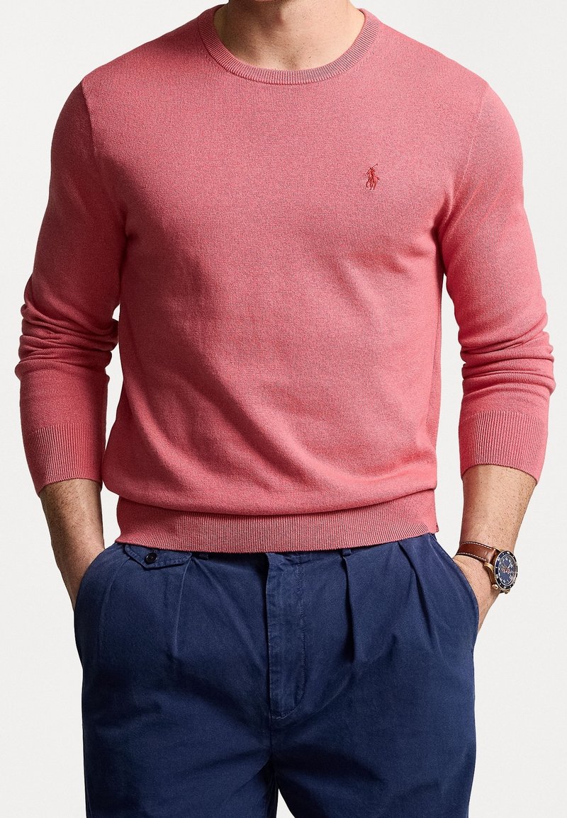 Homme portant un pull ras du cou corail avec un logo brodé, un pantalon bleu marine plissé, et une montre-bracelet avec un bracelet en cuir marron.