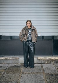 Műszőr barna dzseki egy mintás pulóver fölött, fekete széles szárú nadrággal párosítva. A háttér szürke, texturált.