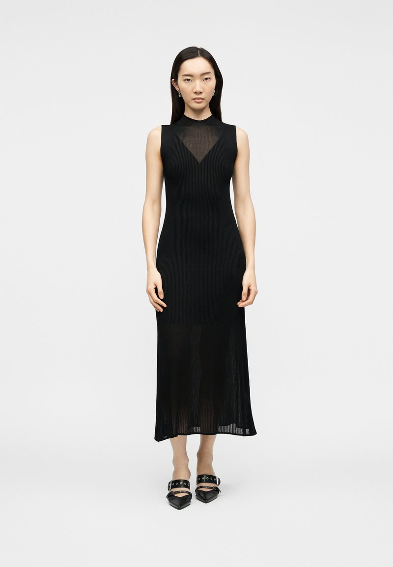 Femme aux cheveux noirs lisses portant une robe midi noire sans manches avec un col transparent et des chaussures noires pointues à brides.