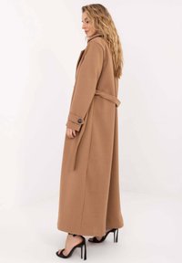 Długi, dopasowany płaszcz z materiału w kolorze camel z gładką fakturą. Posiada pasek w talii, boczne rozcięcia oraz mankiety na guziki.
