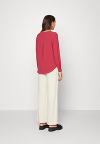 Blusa vermelha de manga longa com uma pregue nas costas, combinada com calças largas em tom creme e sandálias de plataforma pretas. Tecido suave, corte descontraído.