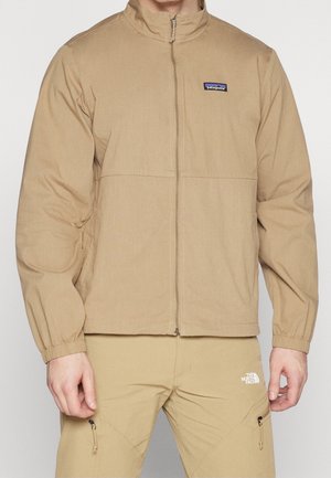 Veste zippée beige Patagonia portée avec un pantalon beige The North Face, vue de face du cou jusqu'à mi-cuisse.