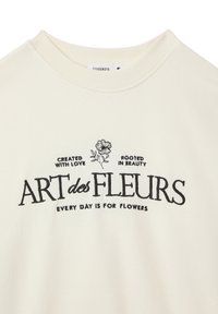 Felpa crema con testo ricamato nero: "ART des FLEURS" con accenti floreali. Collo a girocollo a costine e texture morbida.