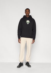 Μαύρο φούτερ με κουκούλα και μπροστινή τσέπη, διακοσμημένο με γραφικό σχέδιο, συνδυασμένο με ανοιχτό μπεζ παντελόνι φόρμας και μαύρα αθλητικά παπούτσια. Απλό, χαλαρό σιλουέτα.