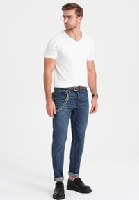 T-shirt bianco a scollo a V, jeans in denim blu scuro con dettagli in catena, maniche arrotolate e scarpe nere con lacci. Sfondo chiaro, in posa in piedi.