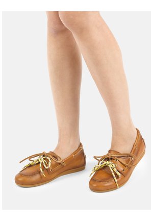 Nogi w brązowych skórzanych butach w stylu mokasynów z żółto-niebieskimi sznurowadłami na białym tle.