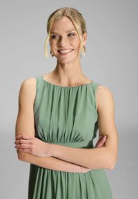 Robe verte sans manches avec une taille froncée, en tissu léger et d'une texture lisse. Modèle avec les bras croisés, fond neutre.