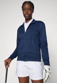 Woman wearing navy zip-up jacket, white polo, and white golf glove holding a golf club against a plain background.