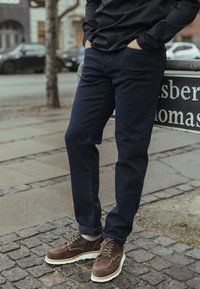 Temno modre ozke kavbojke iz jeansa, kombinirane s rjavimi semiš čevlji z belimi podplati in vezalkami. Vidna stran ulice.