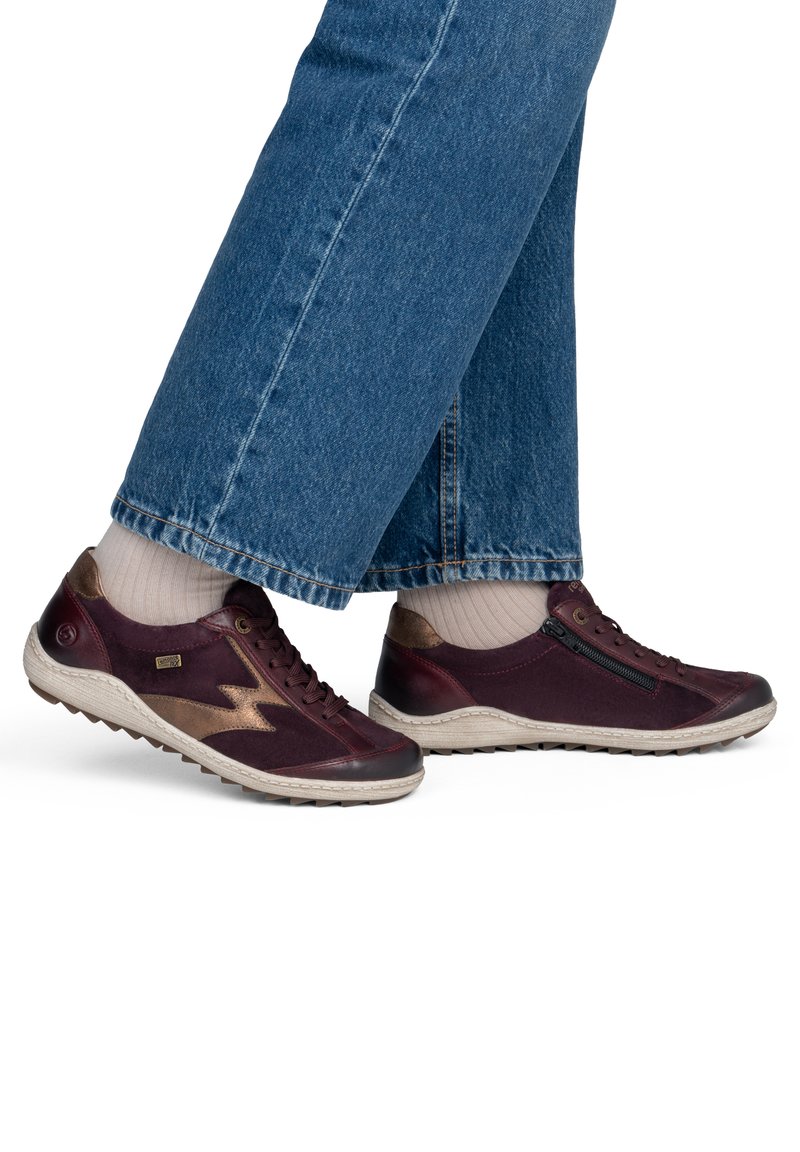 Personne portant un jean bleu, des chaussettes beiges et des baskets décontractées rouge foncé avec des accents beiges et une fermeture éclair latérale sur fond blanc.