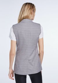 Grå rutmönstrad väst gjord av tyg med en slät textur. Har en skräddarsydd passform och en stilren bakdesign, kombinerad med en vit t-shirt under.