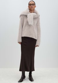 Woman wearing glasses, a fluffy light beige sweater with scarf detail, a long dark brown skirt, and black ankle boots, standing against a plain background.