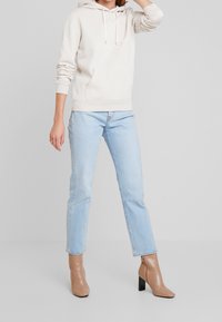 Beige-hupparipaita, jossa on etutasku ja kirjailtu logo, yhdistetty vaaleansinisiin suoralahkeisiin farkkuihin ja ruskeisiin nilkkureihin, joissa on paksu korko.