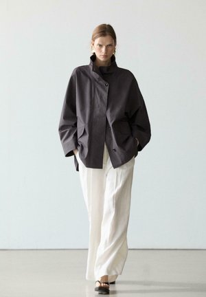 Frau in locker sitzender, dunkel geknöpfter Jacke, weißen weit geschnittenen Hosen, schwarzen offenen High Heels und großen goldenen Ohrringen vor einfarbigem Hintergrund.