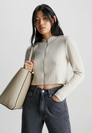 Light beige ribbed cropped top with a high collar and front zipper, paired with dark denim jeans and a beige tote bag with a contrasting edge.