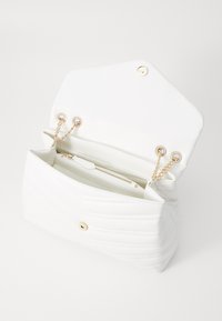Vit quiltad handväska med guldfärgade detaljer, kedjearmar och ett dragkedjefack inuti. Har en distinkt klaffstängning med tryckknappar.