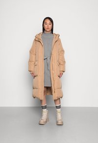 Beige Pufferjacke mit Kapuze, Knopf- und Reißverschluss, zwei Taschen und gesteppter Textur, getragen über einem grauen Kleid und beigen Stiefeln.