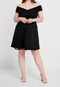 Svart spetsklänning med off-shoulder design, en figurnära bodice och en utsvängd kjol. Bärs med vita klackar med remmar.