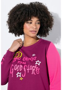 Haut à manches longues en coton prune avec des manches roses, arborant un texte coloré et des graphismes d'étoiles. Texture douce et coupe décontractée.
