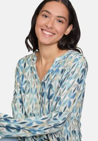 Jeune femme aux cheveux bruns souriant, portant un chemisier à motifs bleus et beiges avec des manches longues et des boutons sur le devant.
