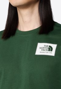 Zbliżenie na osobę w zielonej koszuli z naszywką "The North Face Berkeley, Calif. USA" na klatce piersiowej.