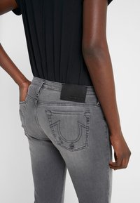 Szare dżinsy o dopasowanym kroju, z czarną skórzaną naszywką na tylnej talii i charakterystycznym przeszyciem na kieszeniach.