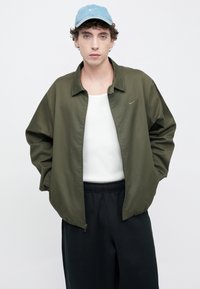 Oļīvu zaļa bombera jaka ar rāvējslēdzēju priekšpusē, asu apkakli un elastīgiem aprocēm, valkāta virs baltas tanktopas un melnām biksēm. Izšūts ar Nike logotipu.