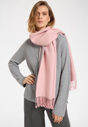 Femme portant un pull et un pantalon gris côtelés, avec une grande écharpe rose à franges enroulée autour du cou, debout avec une main sur la hanche.