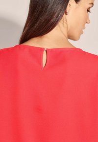 Rote Bluse mit einem Schlüsselloch-Ausschnitt hinten, gefertigt aus einem glatten, leicht glänzenden Material und in einem einfachen, minimalistischen Design.