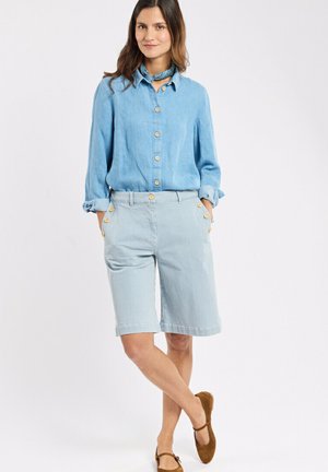 Femme portant une chemise boutonnée bleu clair, un short rayé assorti à boutons jusqu'aux genoux, un foulard bleu autour du cou et des chaussures plates marron.