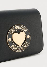 Wallet nero con texture, caratterizzato da un'emblema a forma di cuore dorato con il testo "LOVE MOSCHINO" in un design circolare. Borchie decorative su entrambi i lati.
