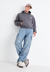 Sweat-shirt à capuche gris avec poche avant, jean cargo bleu clair avec poches latérales, et baskets beige/crème avec accents marron.