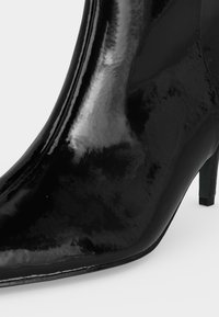 Bottine en cuir verni noir brillant avec bout pointu et talon fin de hauteur moyenne sur fond blanc.