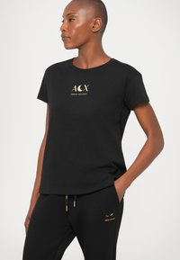 Pessoa vestindo uma t-shirt preta da Armani Exchange e calças pretas combinando, com texto do logótipo em dourado, de pé com uma mão no bolso, virada para a esquerda.