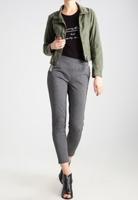 Olivkowa krótka kurtka, czarny t-shirt z grafiką i szare wąskie spodnie w kratę, połączone z czarnymi botkami z odkrytymi palcami.