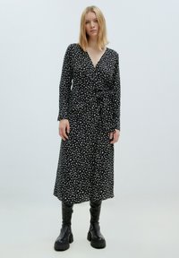 Zwarte wikkeljurk met witte abstracte vlekken, lange mouwen en een geknoopte detail in de taille, gecombineerd met zwarte kniehoge laarzen.