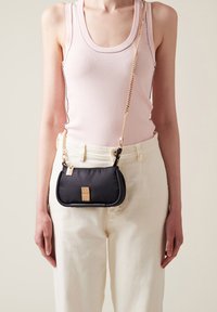 Sac à main noir texturé avec quincaillerie dorée et bandoulière en chaîne, présentant un détail de fermoir rectangulaire, porté par un mannequin avec un pantalon clair.