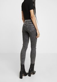 Persona vestida con una camiseta negra de manga corta, jeans skinny de cintura alta en un lavado ácido gris, y botas de tacón negro hasta el tobillo, de pie contra un fondo liso.