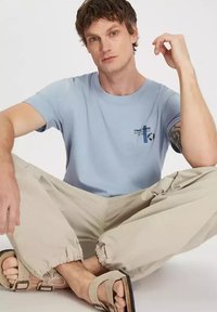 Jasnoniebieska bawełniana koszulka z krótkim rękawem i grafiką z przodu, zestawiona z beżowymi spodniami z elastycznym dołem i jasnymi sandałami.