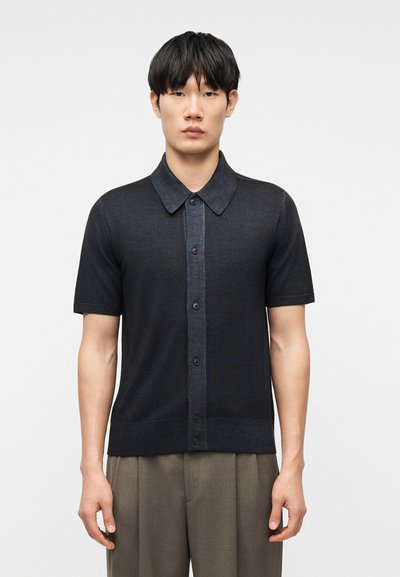 Hombre joven con cabello negro liso, vestido con una camisa oscura de manga corta abotonada y pantalones marrón claro, sobre un fondo liso.