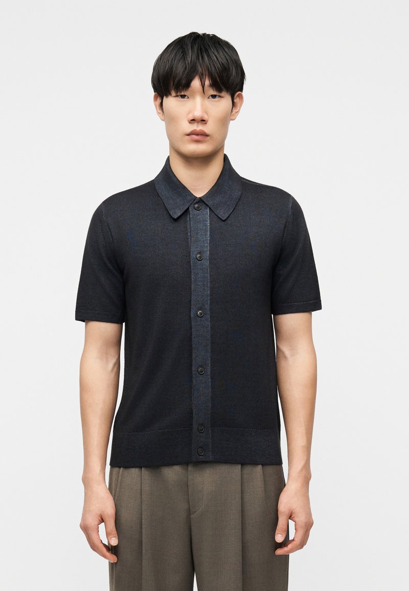 Νέος άνδρας με ίσια μαύρα μαλλιά, φορώντας σκούρο πουκάμισο με κοντά μανίκια και ανοιχτό καφέ παντελόνι, σε ουδέτερο φόντο.