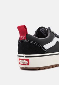 Svart och vit Vans sneakers med röd hälslits och "Vans Off The Wall"-logotyp på vit gummisula, isolerad mot vit bakgrund.