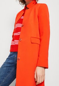 Lys orange frakke med et struktureret design, mandarinhals og fem kontrasterende knapper. Båret over en rødstribet striktrøje og blå jeans.