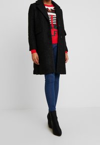 Manteau noir texturé avec col cranté, deux poches avant et boutons, porté par-dessus un pull rouge à motif et un jean skinny bleu. Bottines noires.