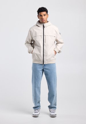 Jonge man staand met handen in de zakken, gekleed in een lichtbeige capuchonjack, lichtblauwe spijkerbroek en witte sneakers, tegen een effen achtergrond.