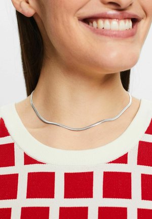 Silberne Choker-Halskette mit einem welligen Design, eng am Hals getragen, kombiniert mit einem rot-weißen gemusterten Pullover. Glatte Textur.